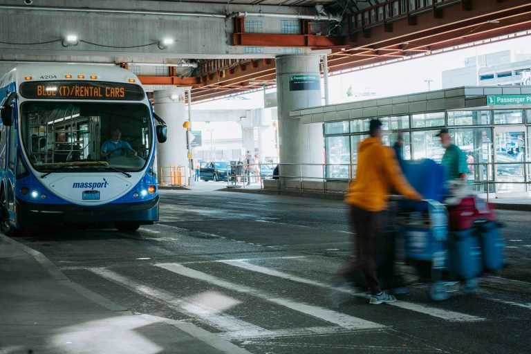 Busy airport scene showing travelers in motion and a rental car bus waiting under a covered terminal.
