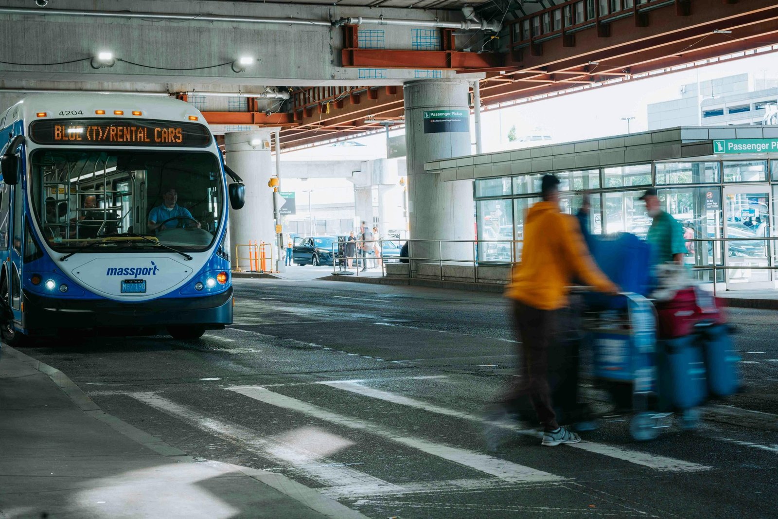 Busy airport scene showing travelers in motion and a rental car bus waiting under a covered terminal.