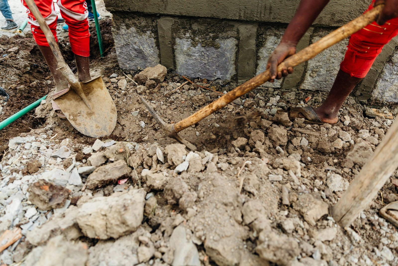 Workers using shovel and pickaxe during construction dig, showing manual labor on rocky ground.