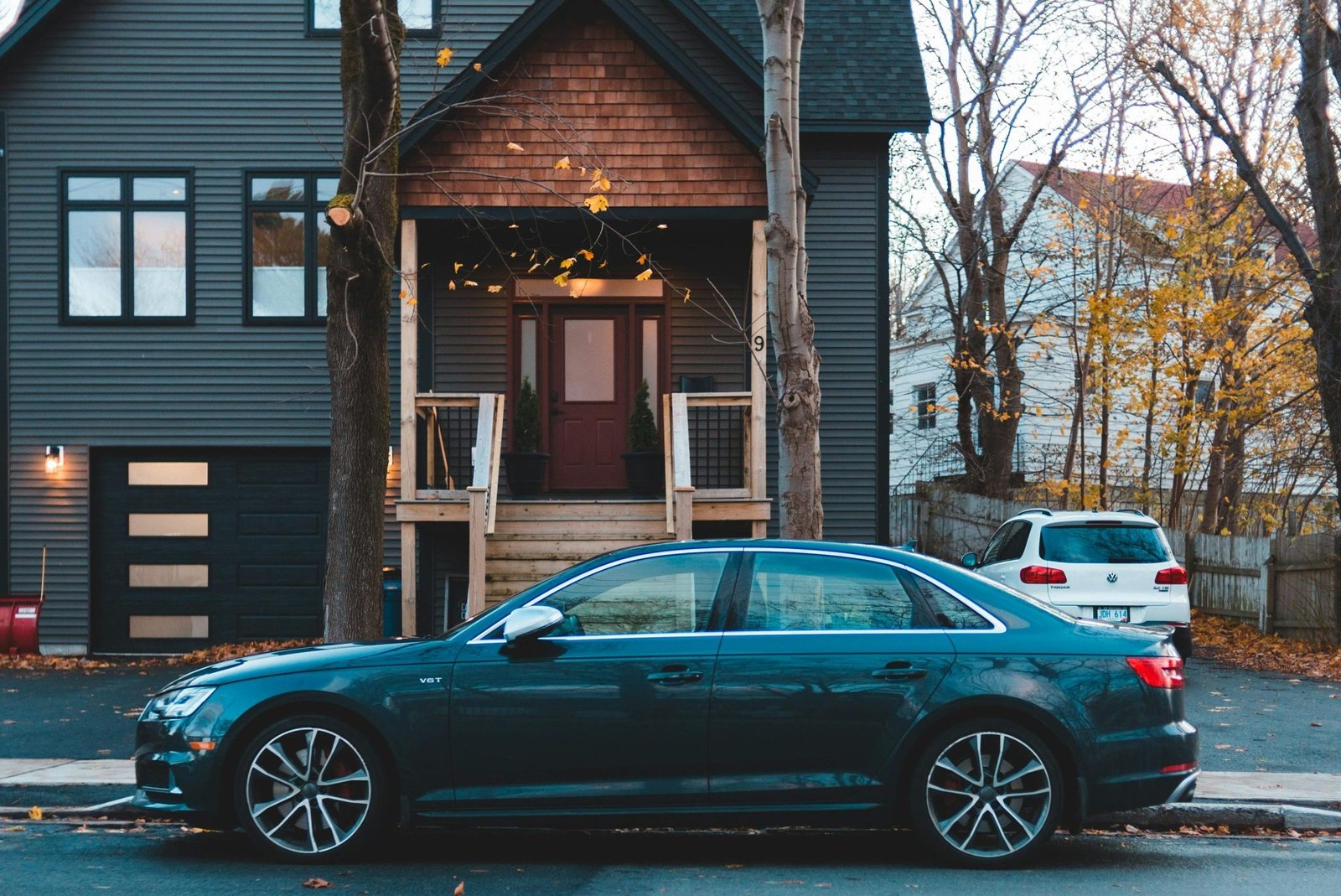 A classic car parked on a street in front of a modern dark-colored house, in an autumn setting.
