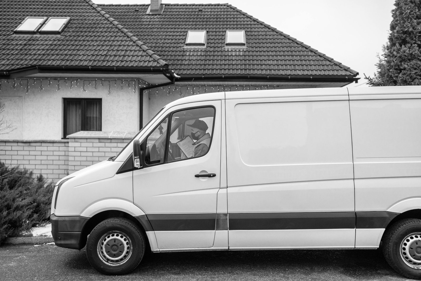 Side view of a white delivery van parked outside a residential house with driver reading a document.