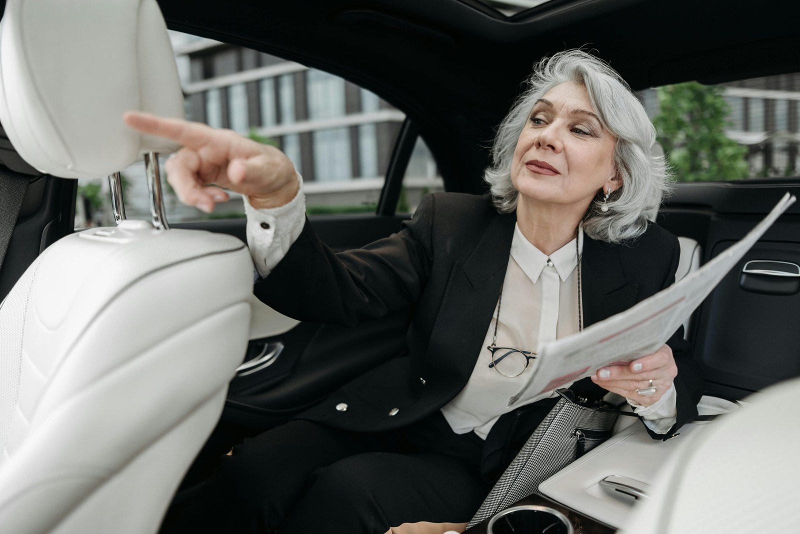 Elegant senior businesswoman in car, pointing with a newspaper in hand, embodying confidence.