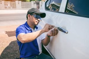 A man wearing sunglasses repairs the door handle of a white car, demonstrating a locksmith's skill in vehicle maintenance.