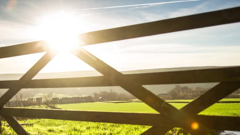 The camera slowly dollys away from a five bar gate, looking though it to a row of houses and a village nestled in a valley beyond a field bathed in bright sunlight.