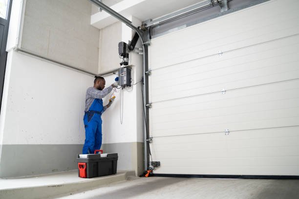 A man in overalls repairs a garage door, focusing on the garage door spring mechanism.