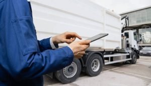 A man in blue overalls holds a tablet next to a truck, promoting online dumpster rental services.