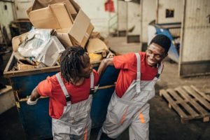 Two men in overalls transporting a cart filled with boxes for a junk removal service.