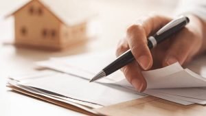 A woman's hand writes on paper with a pen beside a house model, representing mortgage broker services in Gastonia, NC.