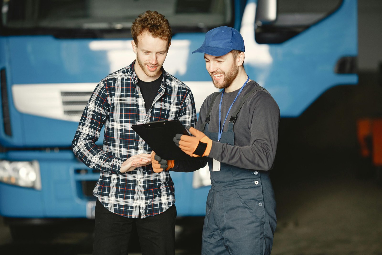 Two men discussing truck repairs in a garage: one in mechanic uniform, the other in a plaid shirt.
