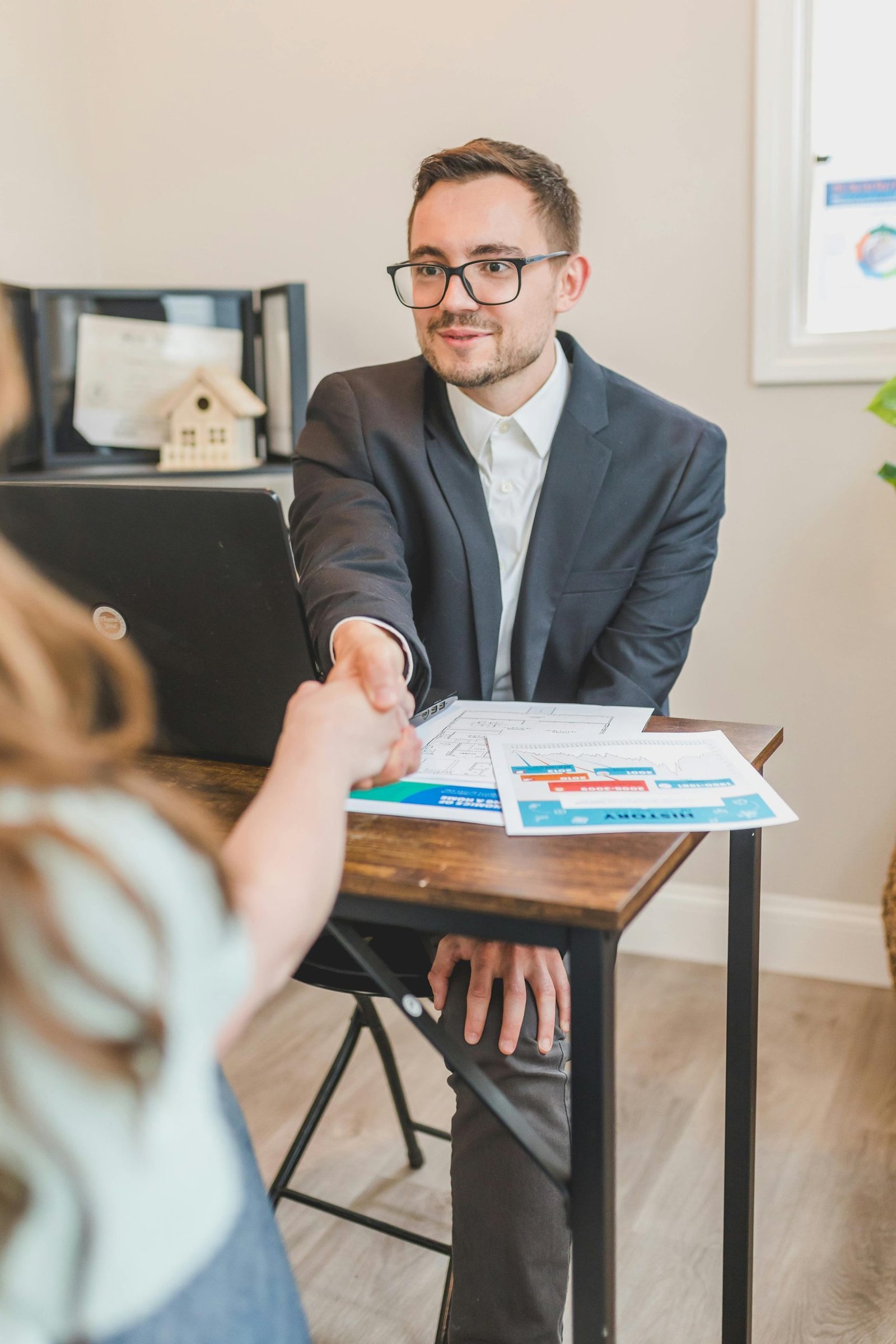 Real estate agent in suit shaking hands with client over property deal papers indoors.