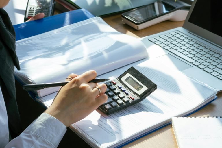 A person working on financial calculations using a calculator and laptop at an office desk.