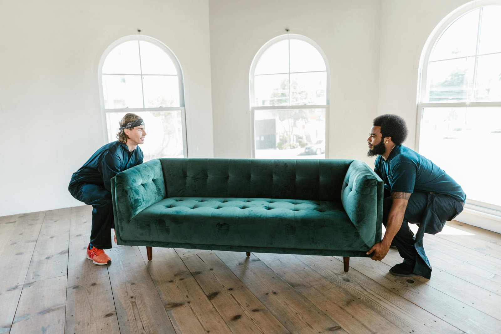 Two men carrying a green sofa in a sunlit room with wooden floors and arched windows.