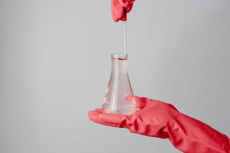 Laboratory scene showing red-gloved hands holding a flask with clear liquid.