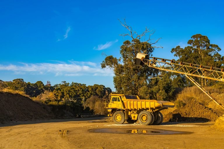A large dump truck and conveyor in a quarry under a bright blue sky.